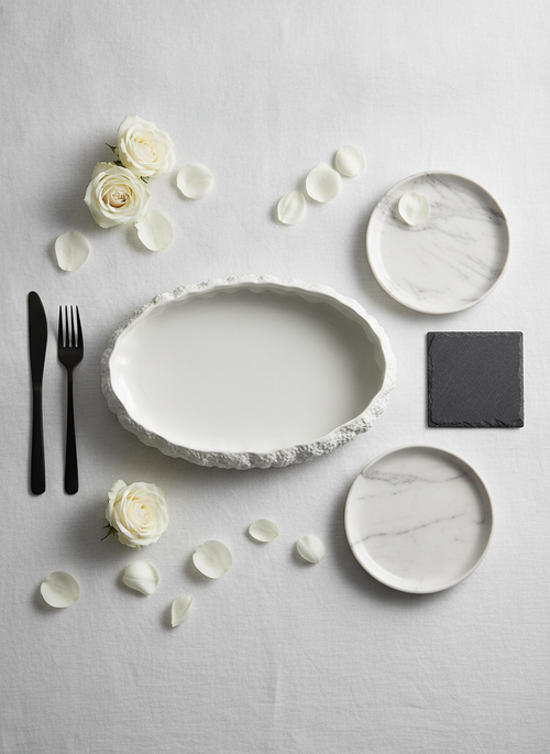 White rock-textured oval porcelain serving platter, editorial flat-lay place setting with matte-black cutlery and white roses