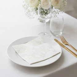 Square gold and white floral cocktail napkin folded flat on a white plate in an elegant table setting with gold cutlery and white roses