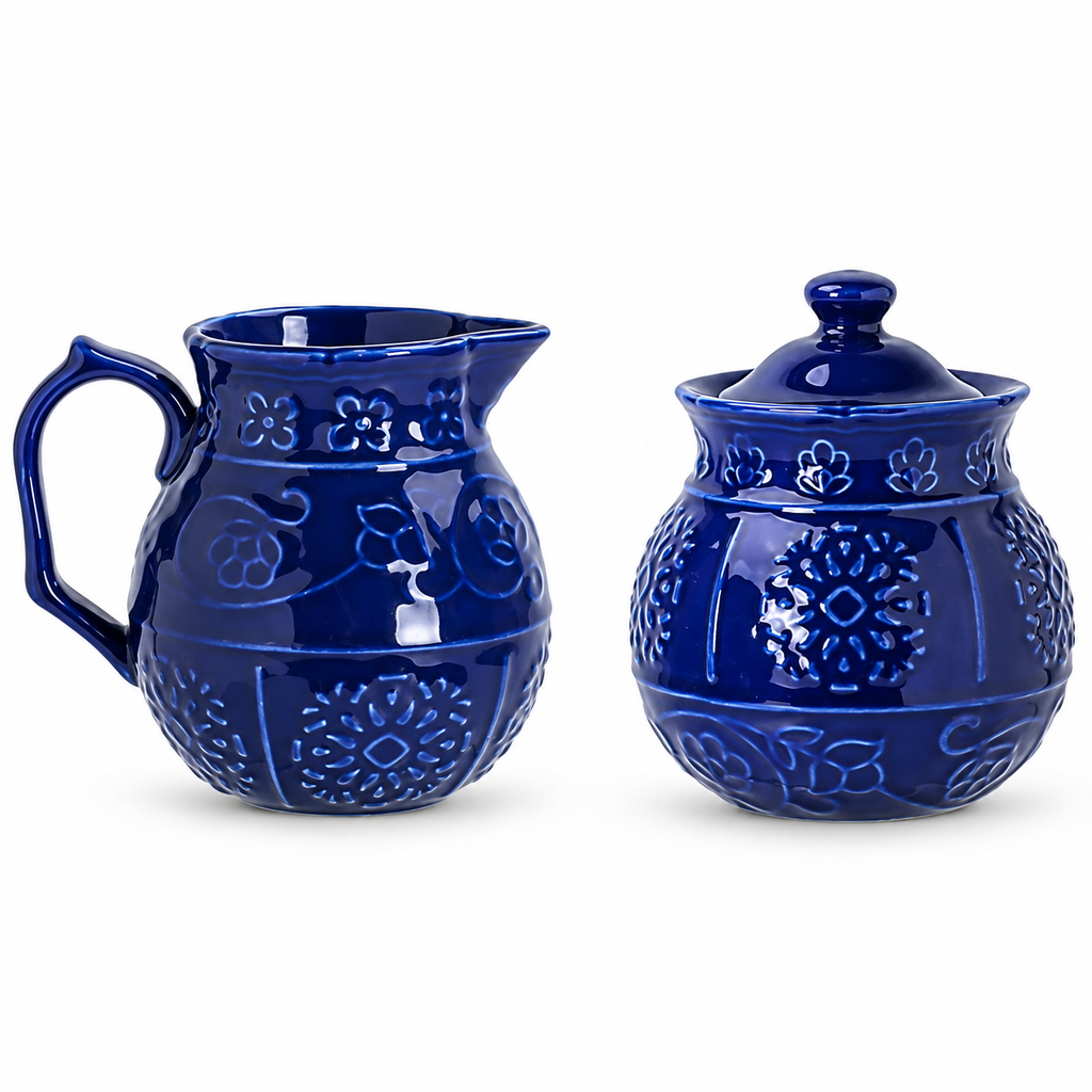 Glossy cobalt-blue embossed ceramic creamer and closed sugar bowl on white, showing the handle, pouring lip, floral relief and fitted lid. Spoon is not shown.