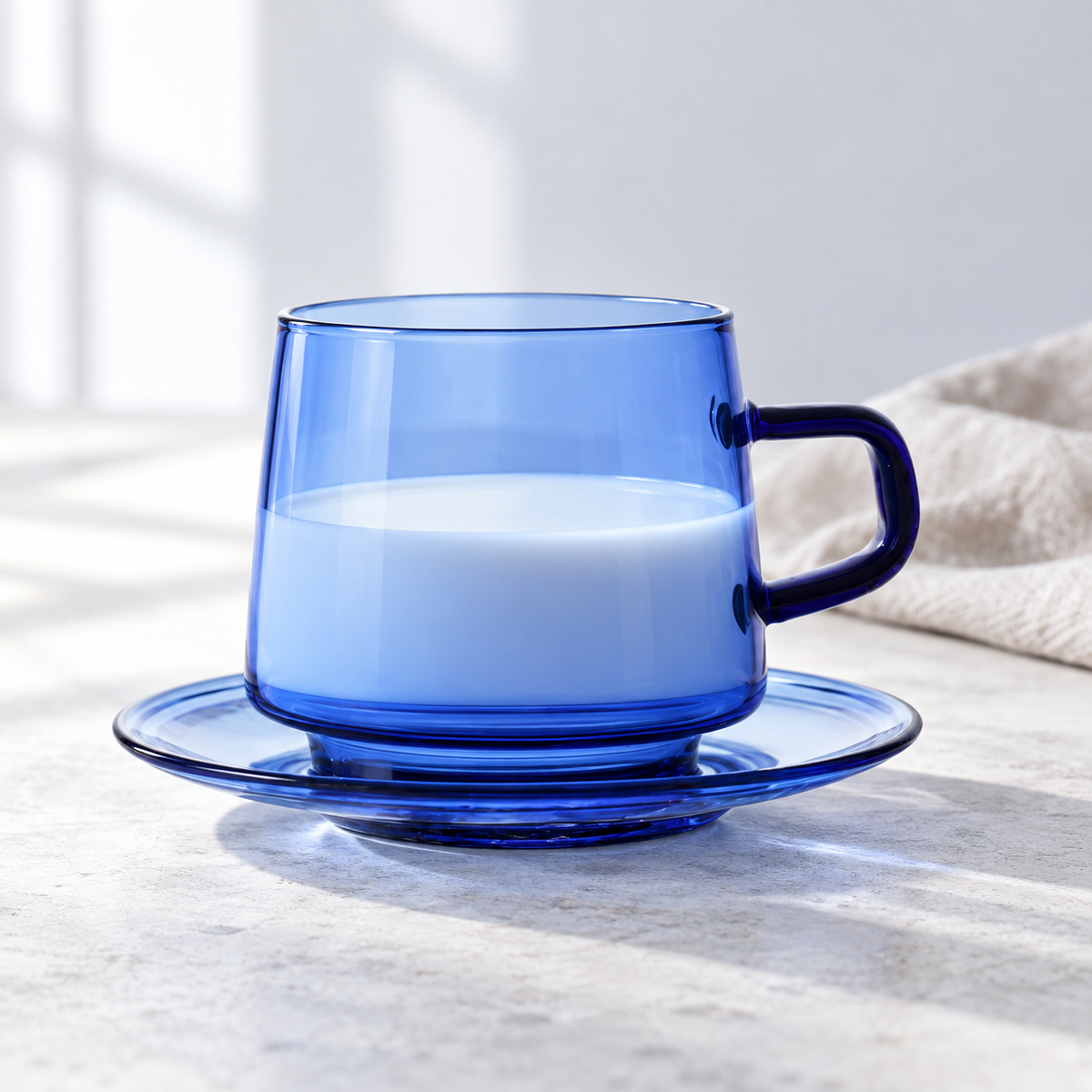 Blue glass cup and matching saucer with milk in a bright setting; beverage and linen are not included.