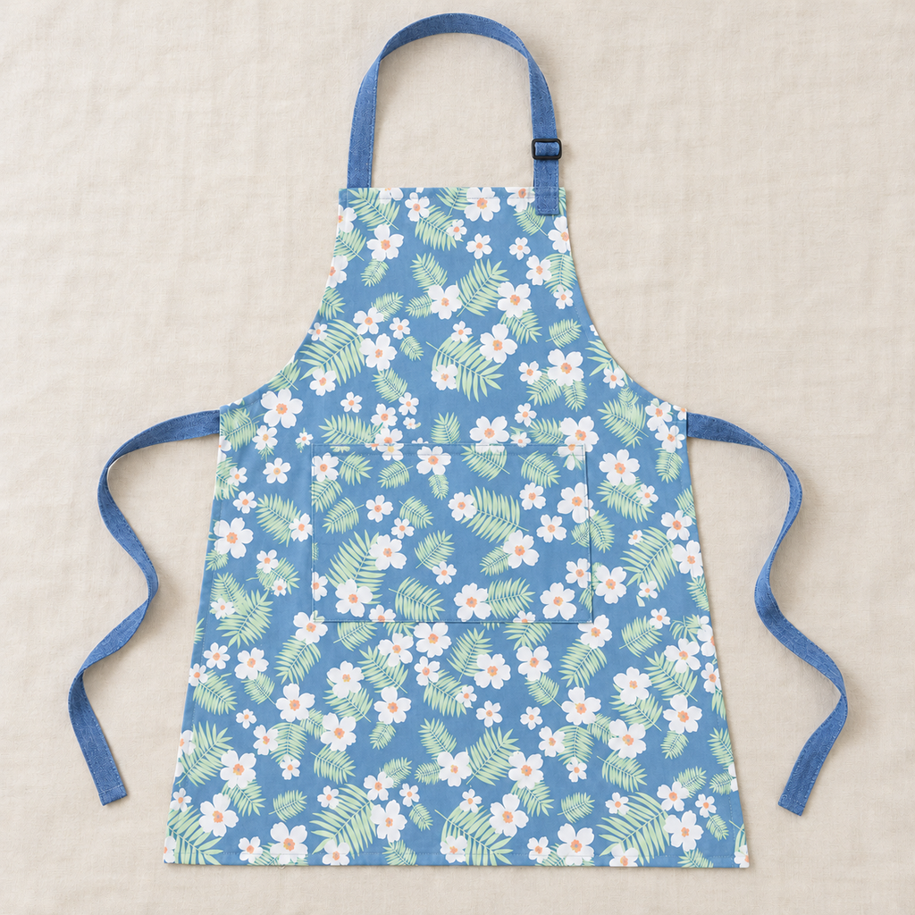 Blue apron with white flowers and green leaves laid flat, showing neck strap, waist ties and rectangular front pocket.