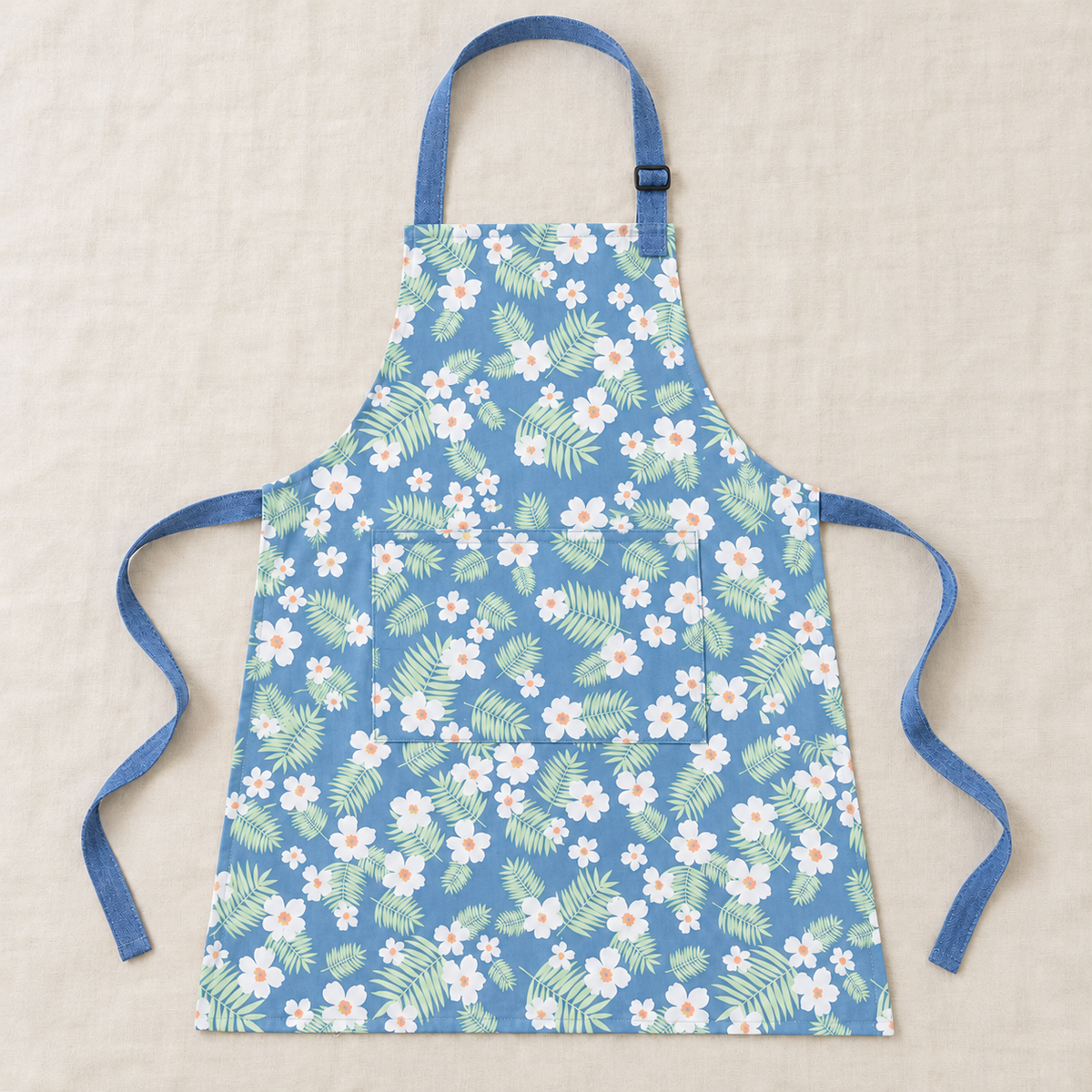Blue apron with white flowers and green leaves laid flat, showing neck strap, waist ties and rectangular front pocket.