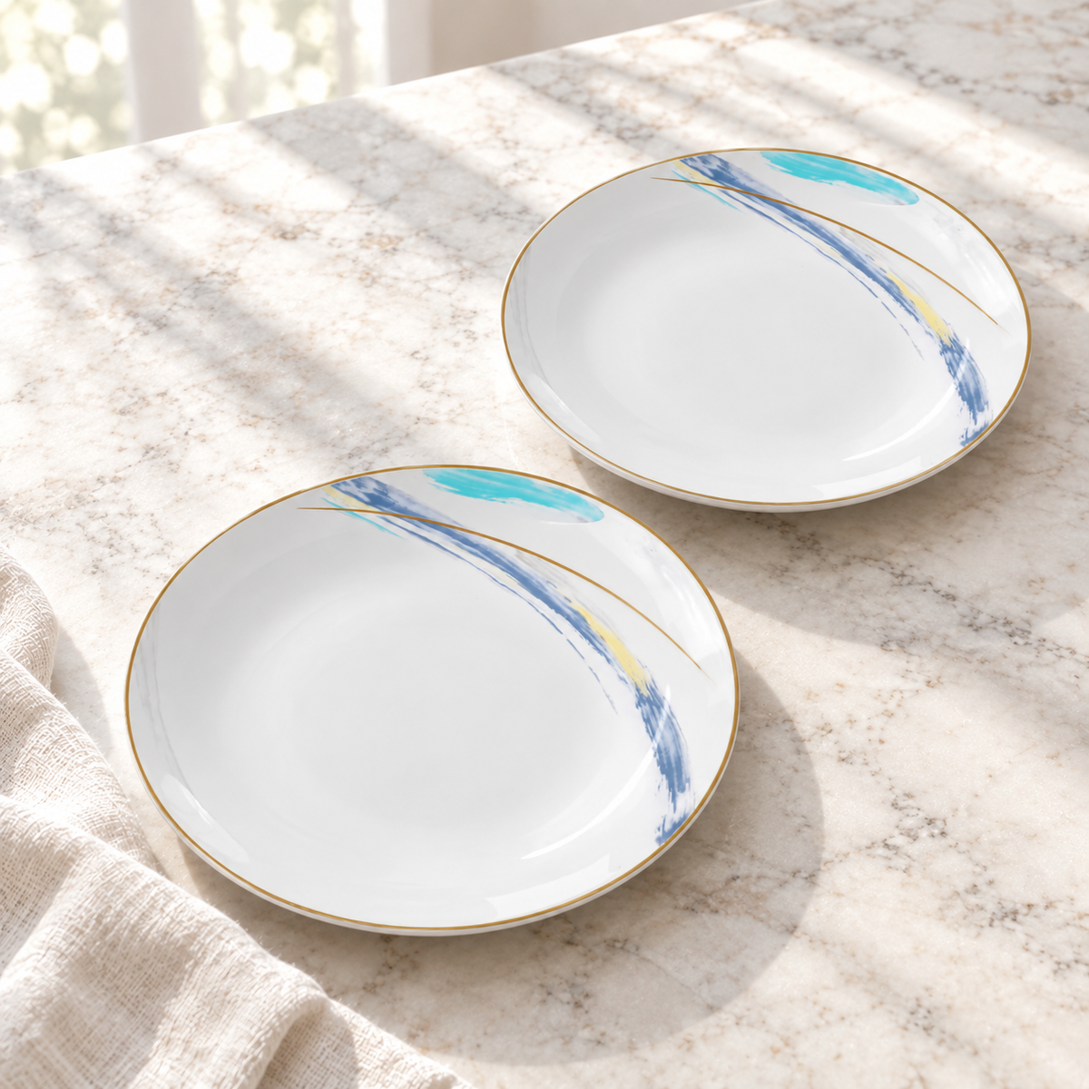 Two Gilded Edge ceramic dessert plates face up on a pale table in natural daylight. Linen is a styling prop. Representative arrangement from the set.