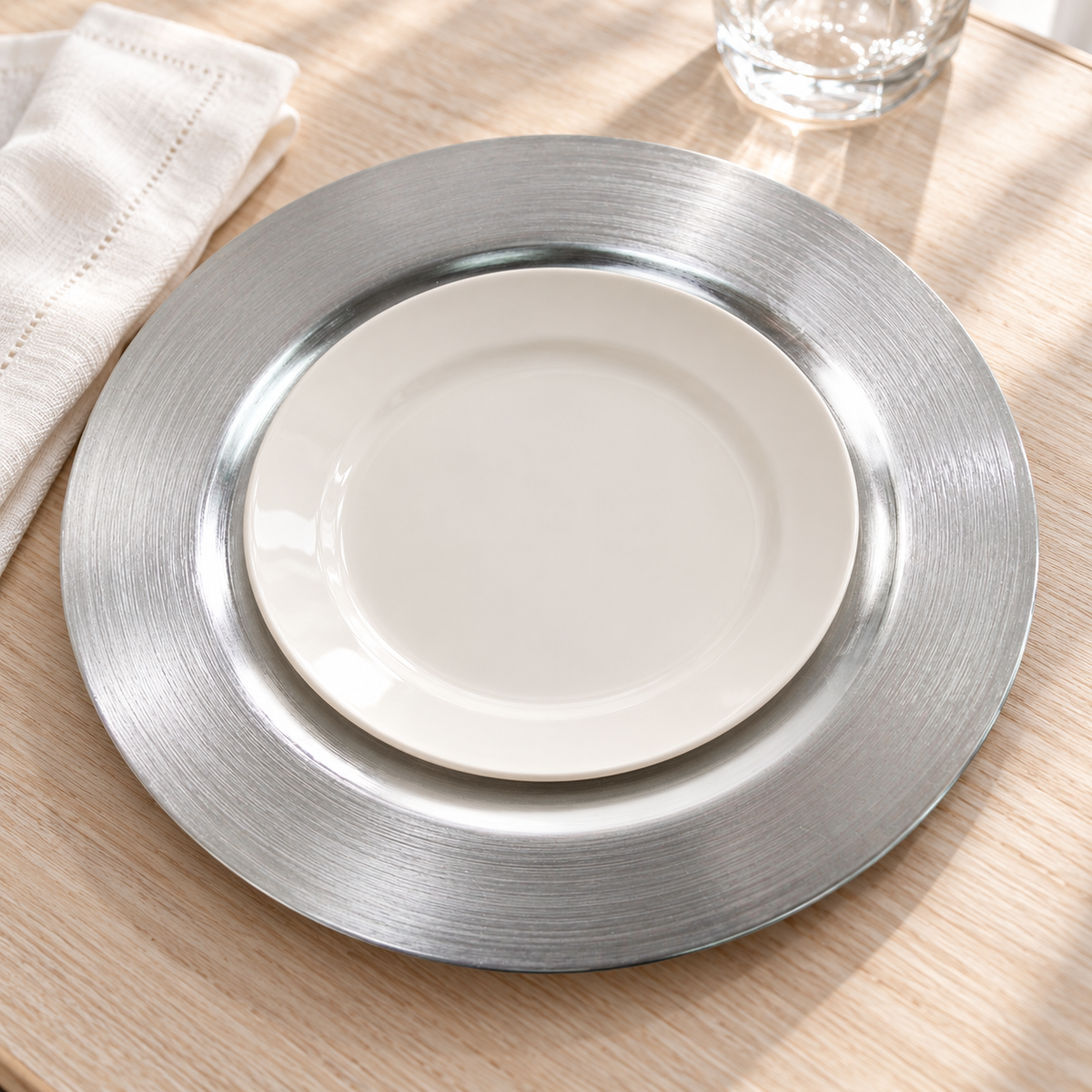 Close view of a metallic silver decorative charger beneath a separate empty ivory plate on pale wood. Styling props not included.