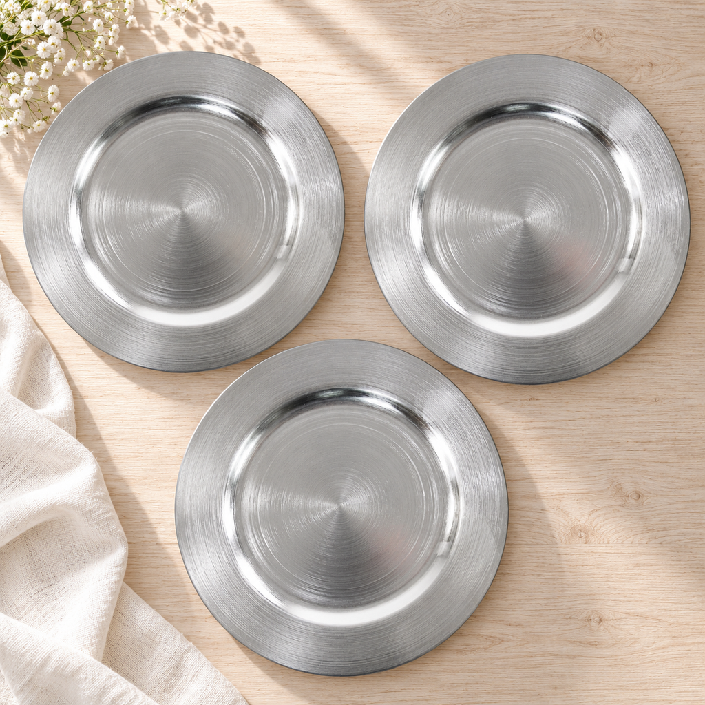 Overhead view of empty metallic silver decorative chargers with fine concentric texture on a pale wood table. Styling props not included.