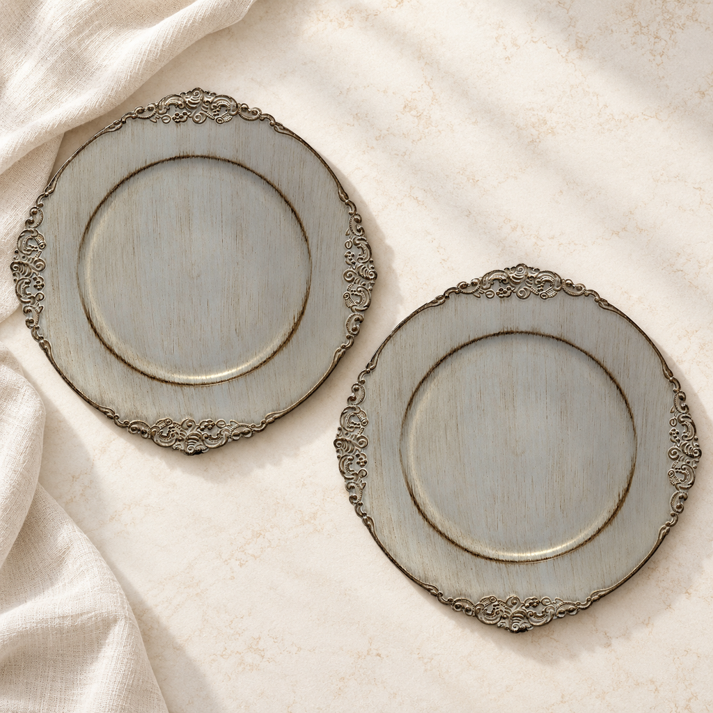 Two empty face-up chargers showing antique grey decorative charger with distressed finish and ornate scroll rim. Representative arrangement from the set.