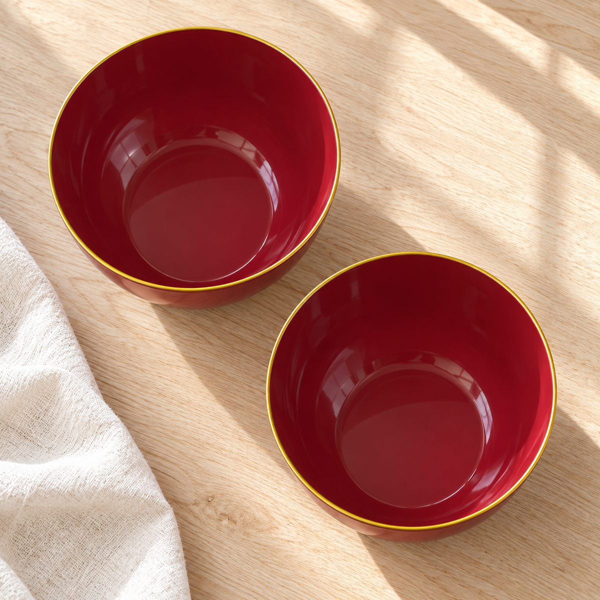 Overhead view of empty burgundy plastic bowls with fine gold rims on pale wood. Food and styling props not included.