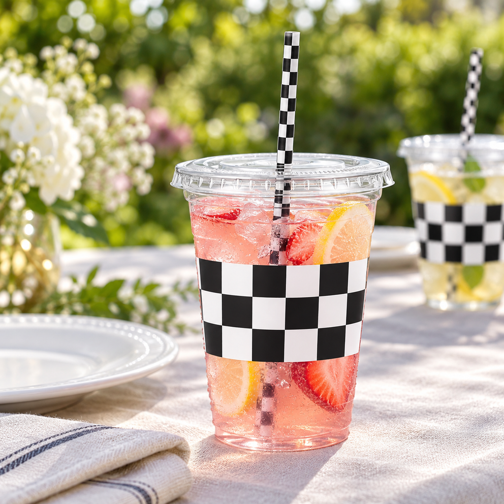 Clear 16 oz checkered party cup with its flat clear lid and checked paper straw, picnic photograph. Representative items from 50 complete sets; drinks and styling props are not included.