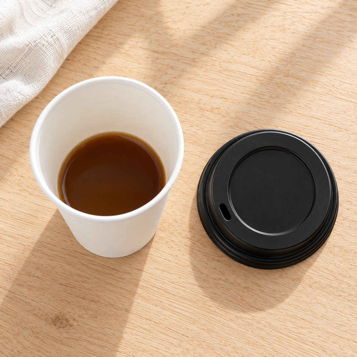 High-angle view of a white paper cup and black sipping lid on a pale wood table. Beverages and styling props not included.