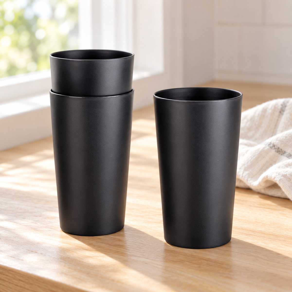 A nested pair of matte black tumblers beside a single matching cup on a light wood counter.