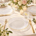 Clear hammered gold rim plates at separate place settings on ivory linen with pale flowers, candles and gold flatware. Representative arrangement; food and styling props not included.