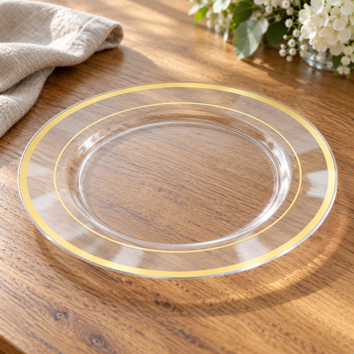 Clear 10-inch plastic plate on oak, showing its thin edge, broad clear sloped rim, wide outer gold band and thin inner gold ring. Food and styling props not included.