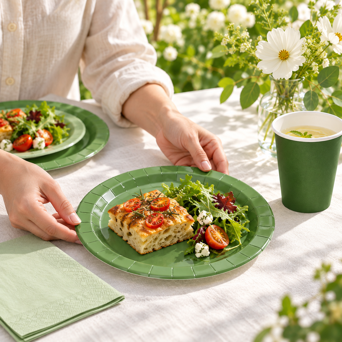 Guest placing a green paper plate with focaccia and salad on an ivory linen garden table beside a plain green paper cup and folded napkin. Representative arrangement; food and styling props not included.
