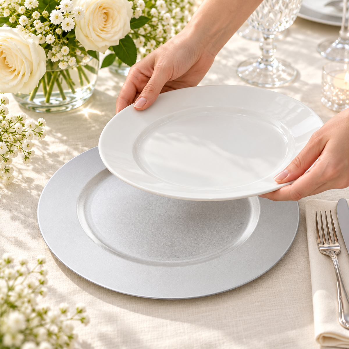 Hands lower a separate white dinner plate toward a silver white charger beside white flowers and table linens. Representative arrangement; food and styling props not included.