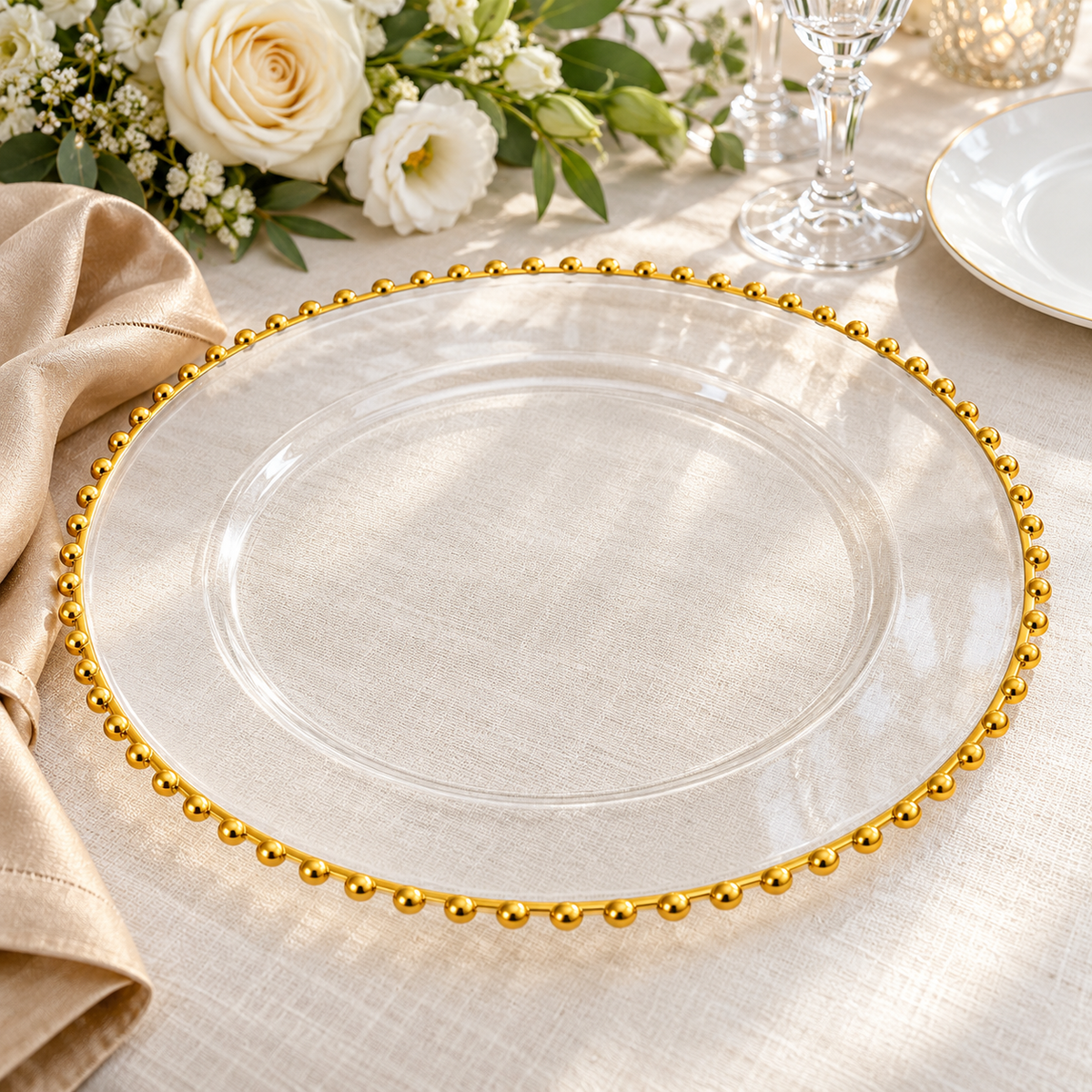 Empty clear charger showing its transparent center, broad smooth rim and single gold bead border on ivory linen beside cream flowers. Representative arrangement; food and styling props not included.