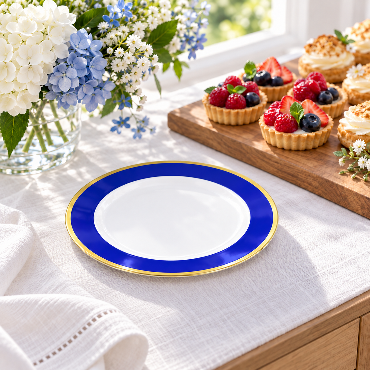 A single small blue and gold plate beside fruit tartlets on a wooden board at a white linen dessert buffet. Representative arrangement; food and styling props not included.