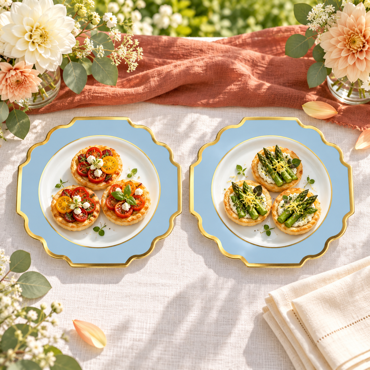 Two separate white, dusty blue and gold small plates holding tomato and asparagus tartlets on ivory linen with a terracotta runner and dahlias. Representative arrangement; food and styling props not included.
