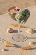 Green shell goblet holding a pale spritz cocktail on a travertine table with small seashells
