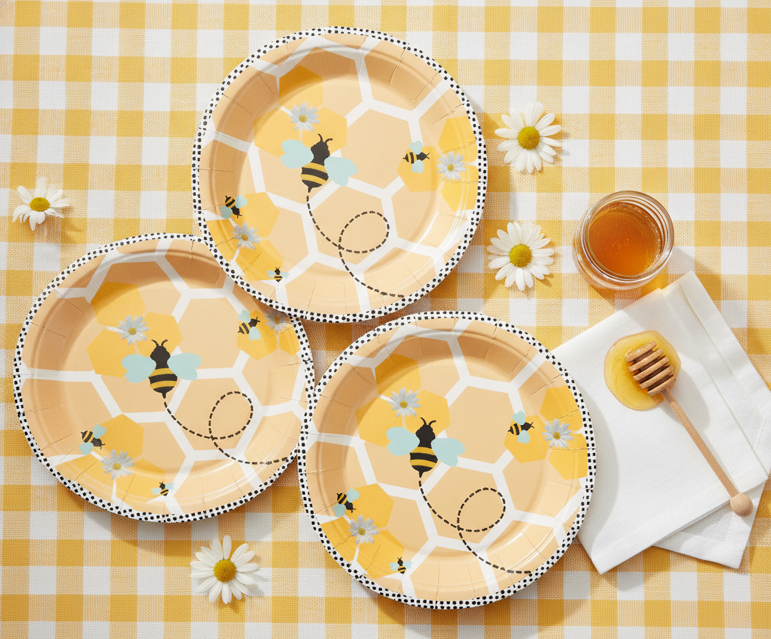 Overhead flat-lay of bee-design paper plates with honey and daisies on gingham