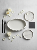 White rock-textured oval porcelain serving platter, editorial flat-lay place setting with matte-black cutlery and white roses