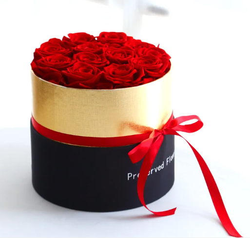 Round gift box packed with red preserved roses, gold band over a black base and a red ribbon bow