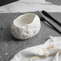 Deep rock-textured white oval bowl, three-quarter hero on marble and slate