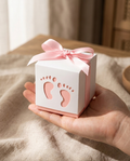 Hand holding a white baby footprint favor box with pink ribbon