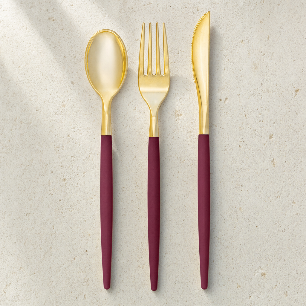 Two-tone gold and cranberry plastic spoon, fork and knife laid side by side on a speckled surface