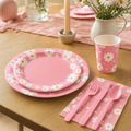 Pink daisy paper plates, cup and pink cutlery laid on a warm oak table with daisies in a stoneware jug