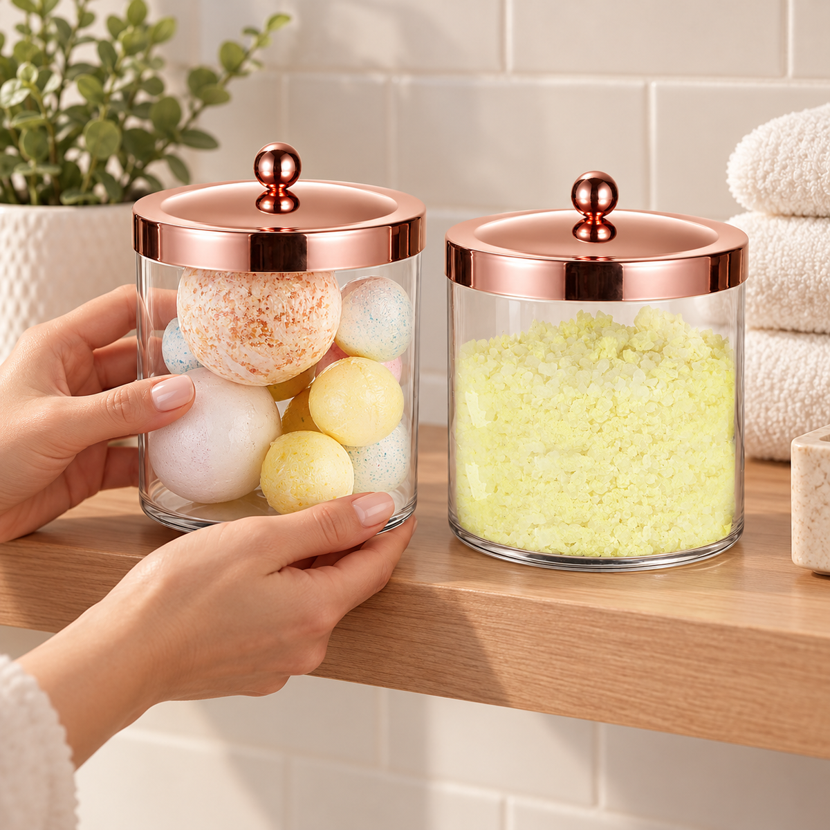 Two closed rose-gold-lid jars arranged on a bathroom shelf; bath contents, hands, towels, plant and shelf are styling props, not included.