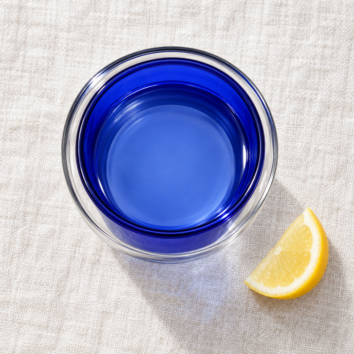 Overhead view of Blue-250mL double-wall glass with water on linen. Lemon is styling only.