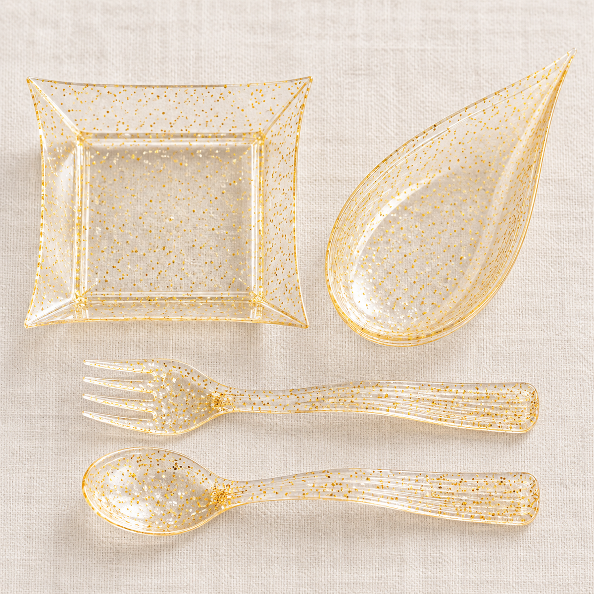 Overhead view of empty clear gold-flecked square and teardrop mini dishes with matching miniature fork and spoon. Linen is a styling prop.