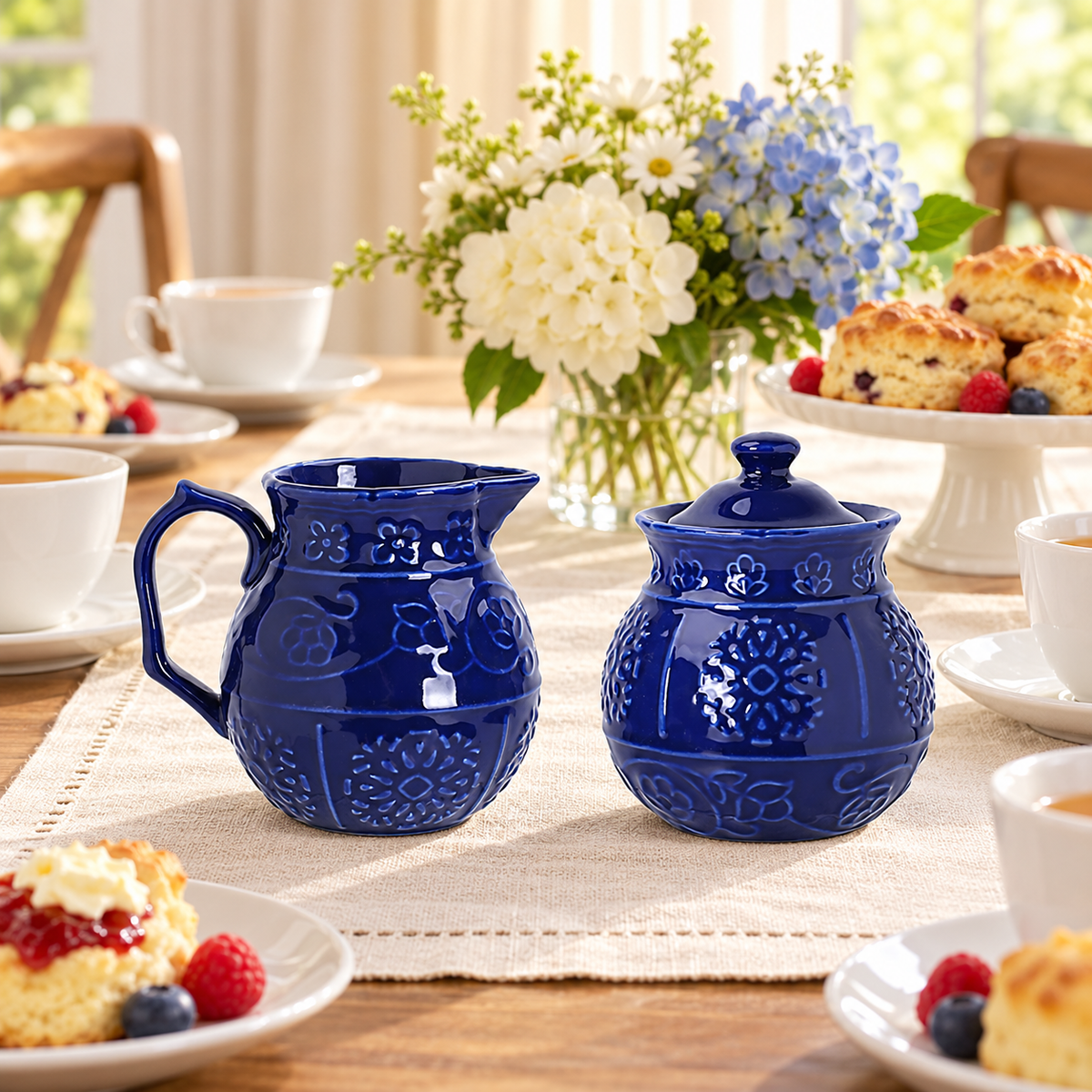 Blue embossed ceramic creamer and closed sugar bowl styled on an afternoon tea table. Other tableware and decor are styling props, not included.