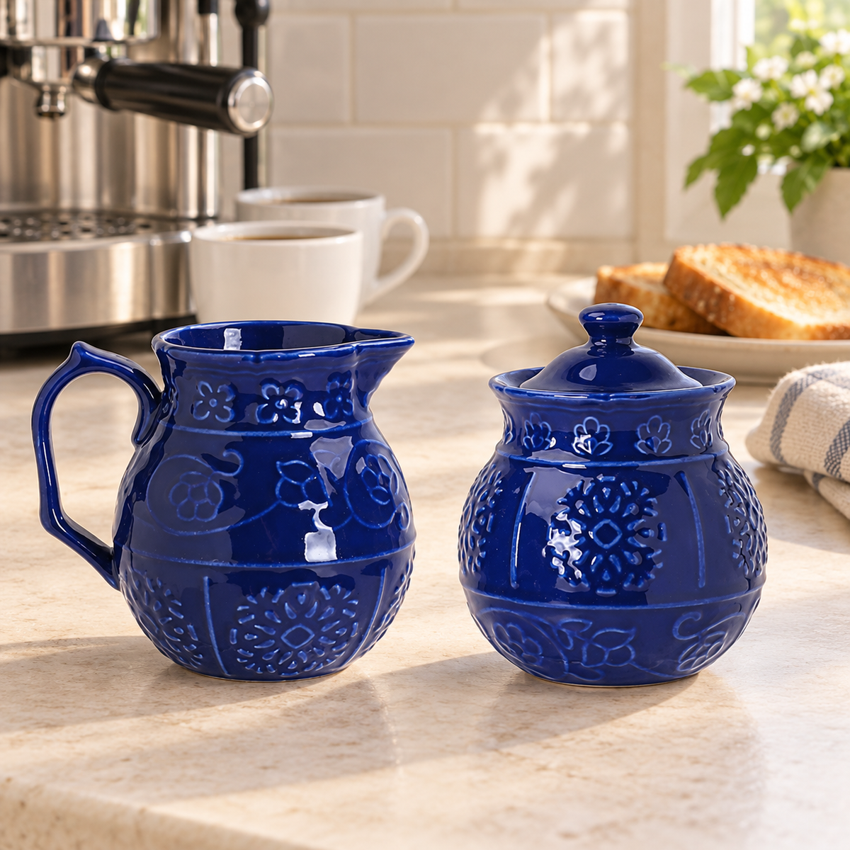 Blue embossed ceramic creamer and closed sugar bowl on a kitchen coffee counter. Coffee equipment and decor are styling props, not included.