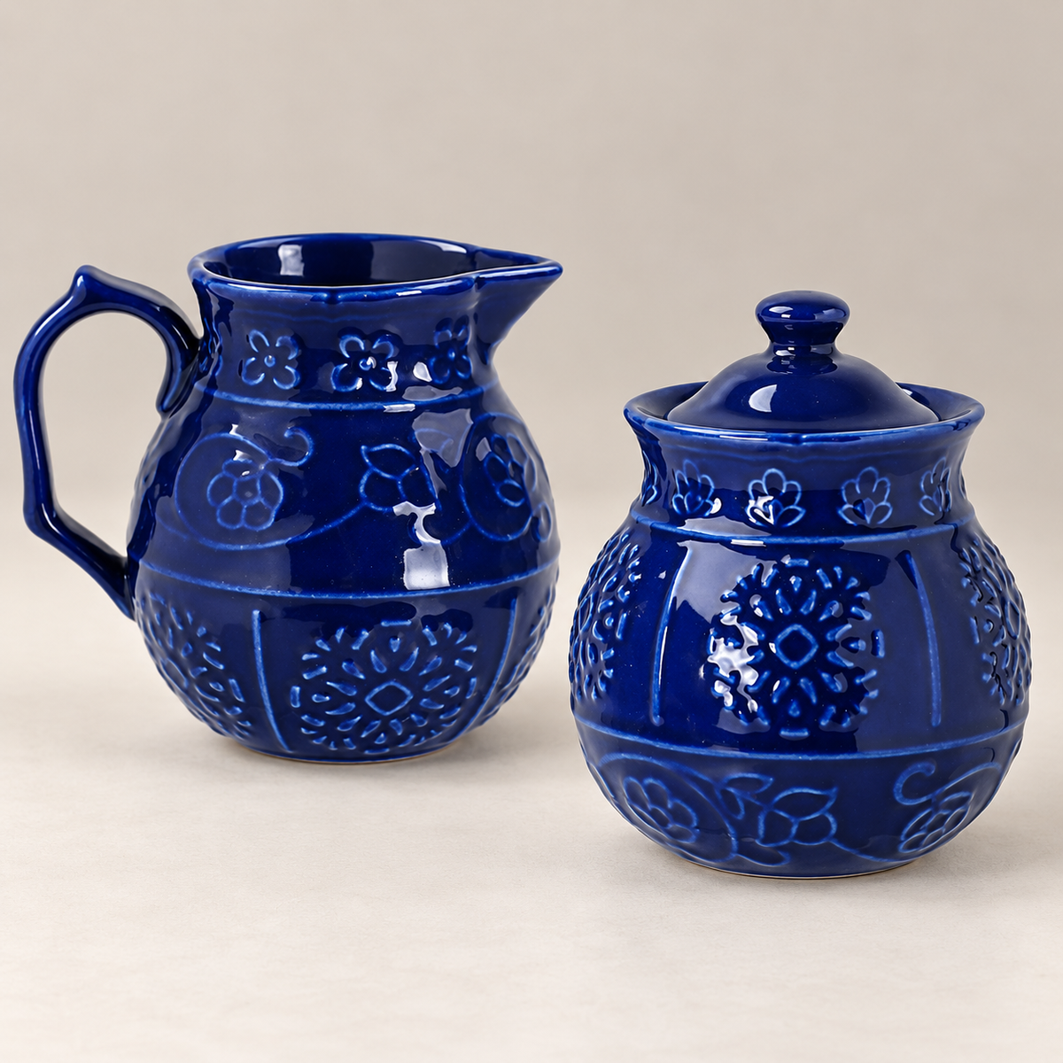 Close-up of the blue ceramic creamer and closed sugar bowl, showing glossy cobalt glaze, raised floral decoration, creamer handle and spout.