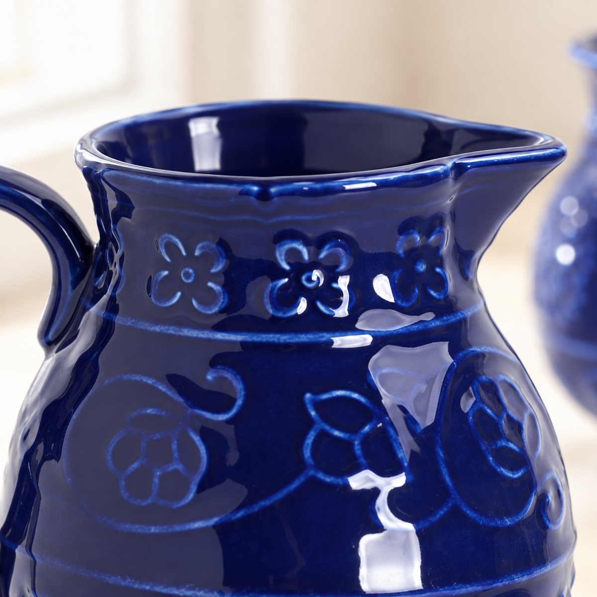 Cropped close-up of the blue ceramic creamer from the sugar-and-creamer set, showing the glossy cobalt glaze, floral relief, rim, spout and part of the handle.