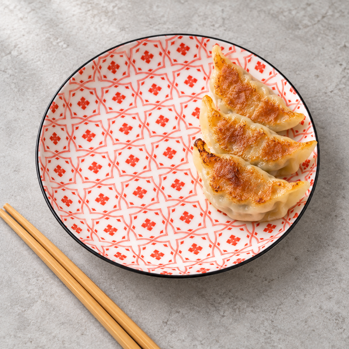 The red geometric-patterned ceramic plate with a black rim serving gyoza. Food and chopsticks are styling props, not included.
