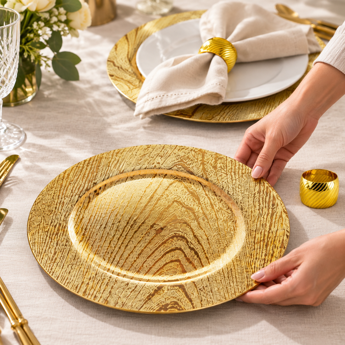 Hands arranging a representative gold woodgrain charger face-up beside its ring and a second place setting. White dinner plate, napkin, cutlery, glassware and decor are props, not included.