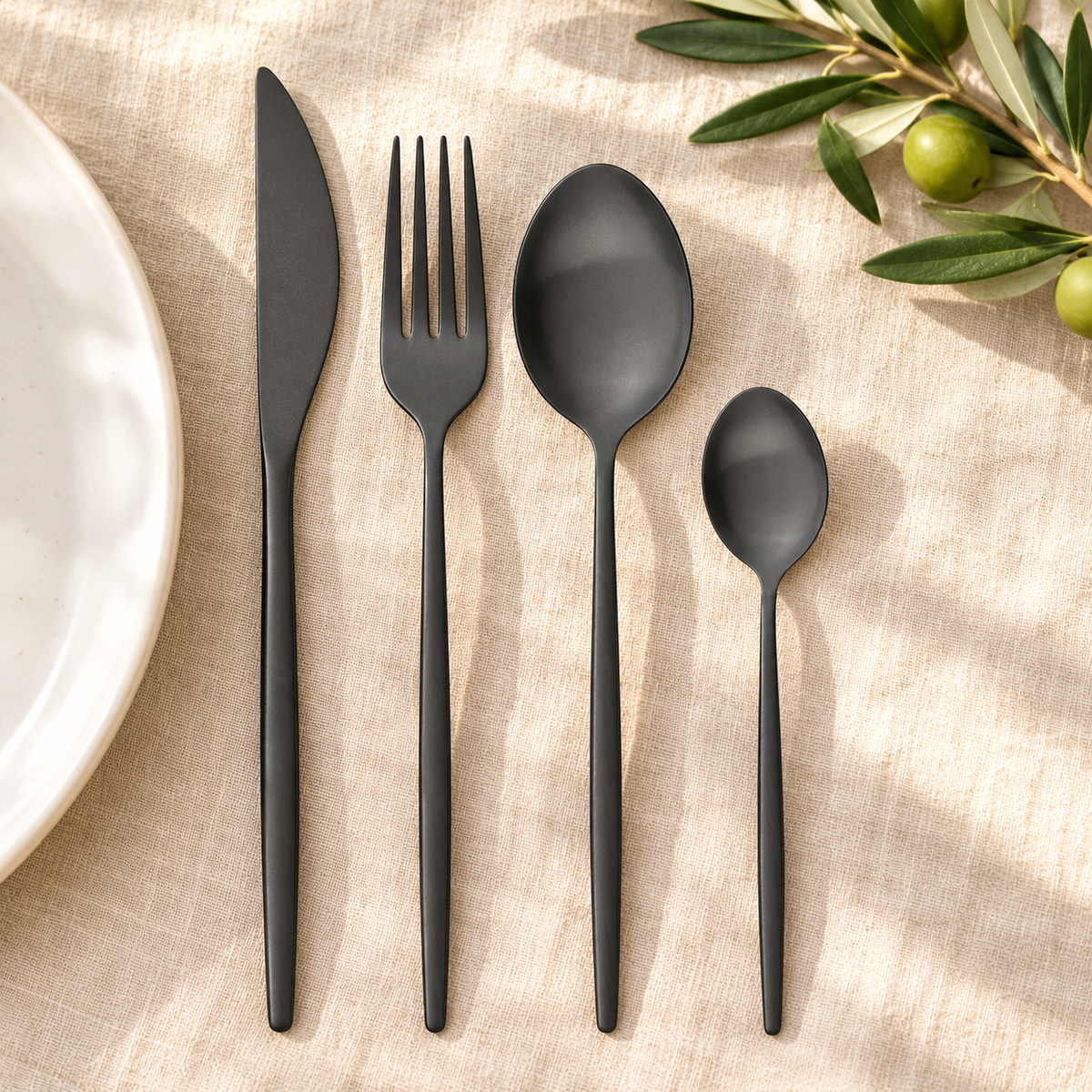 One four-piece matte black cutlery setting on natural linen; plate and olive branch are styling props.