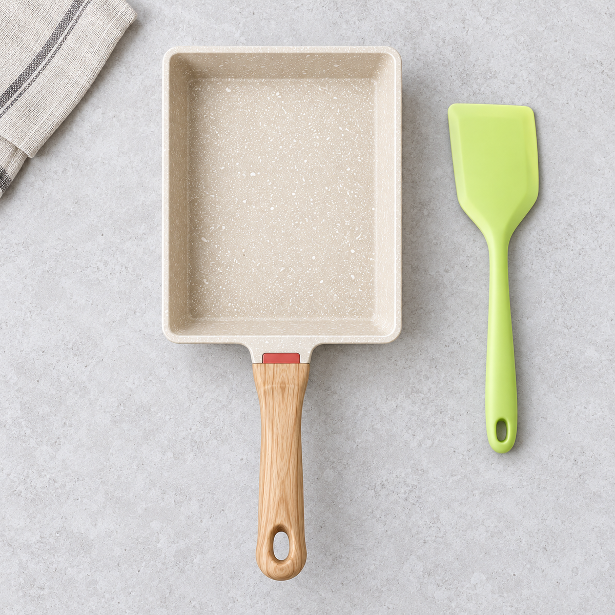 Straight overhead view of empty white speckled rectangular tamagoyaki pan with wood-look handle and supplied green spatula on pale stone; linen not included.