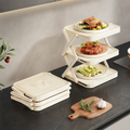 Cream-white foldable 3-tier dish and plate serving rack holding three plates of prepared food on a kitchen counter